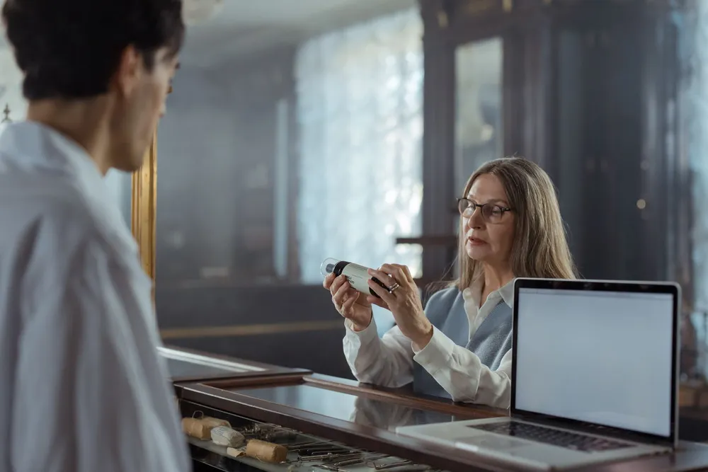 A woman in her fifties with glasses reading the label on a medicine bottle at a counter.